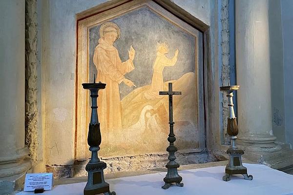 Religious fresco in a church, depicting a saint in prayer, with three candlesticks and a cross in the foreground.