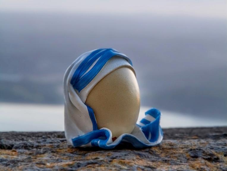 Egg decorated with a white and blue veil, inspired by Mother Teresa’s iconic habit, placed on a rock at sunset.