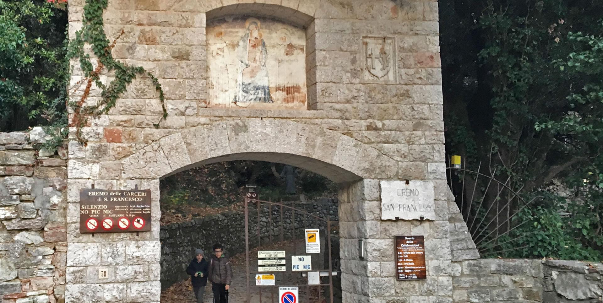 Entrance of the Eremo delle Carceri: a stone arch with a faded sacred fresco, coats of arms, and signs inviting silence.