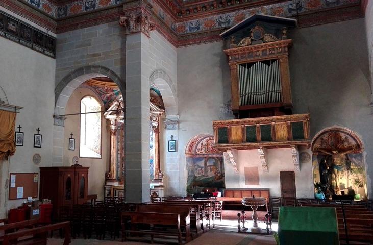 View of the central nave with benches for the faithful and a holy water font. On the sides are confessionals, while in the background stand a crucifix and the organ.