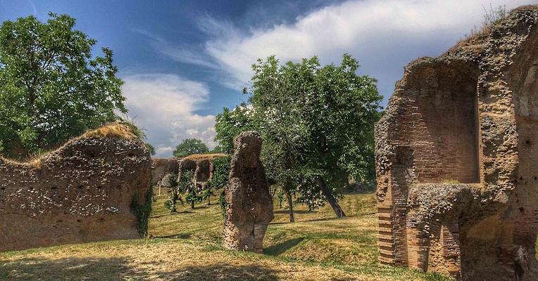 Resti di strutture romane su un prato verde, con alberi sullo sfondo e cielo azzurro attraversato da nuvole.