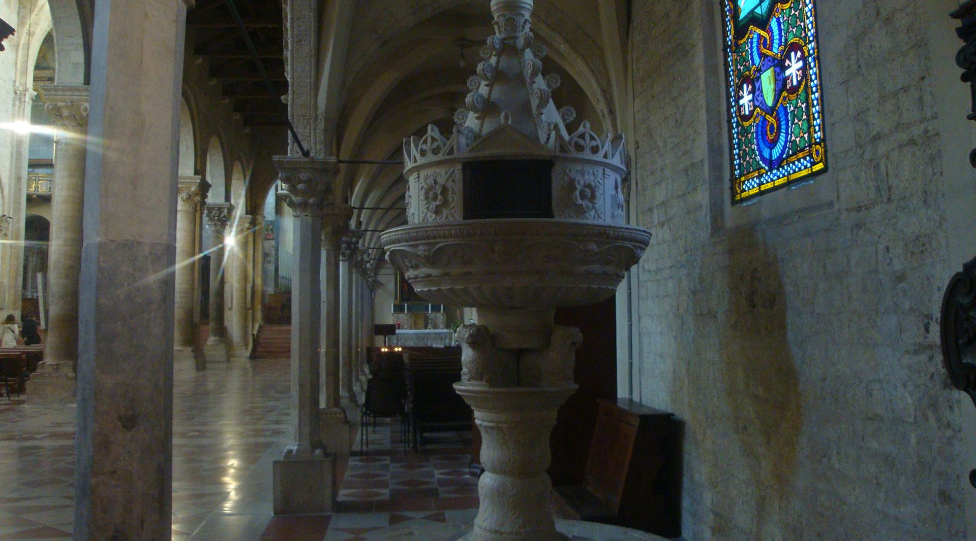 Stone-carved baptismal font in Todi Cathedral, with Gothic details, arched columns, and a decorated stained-glass window in the background.