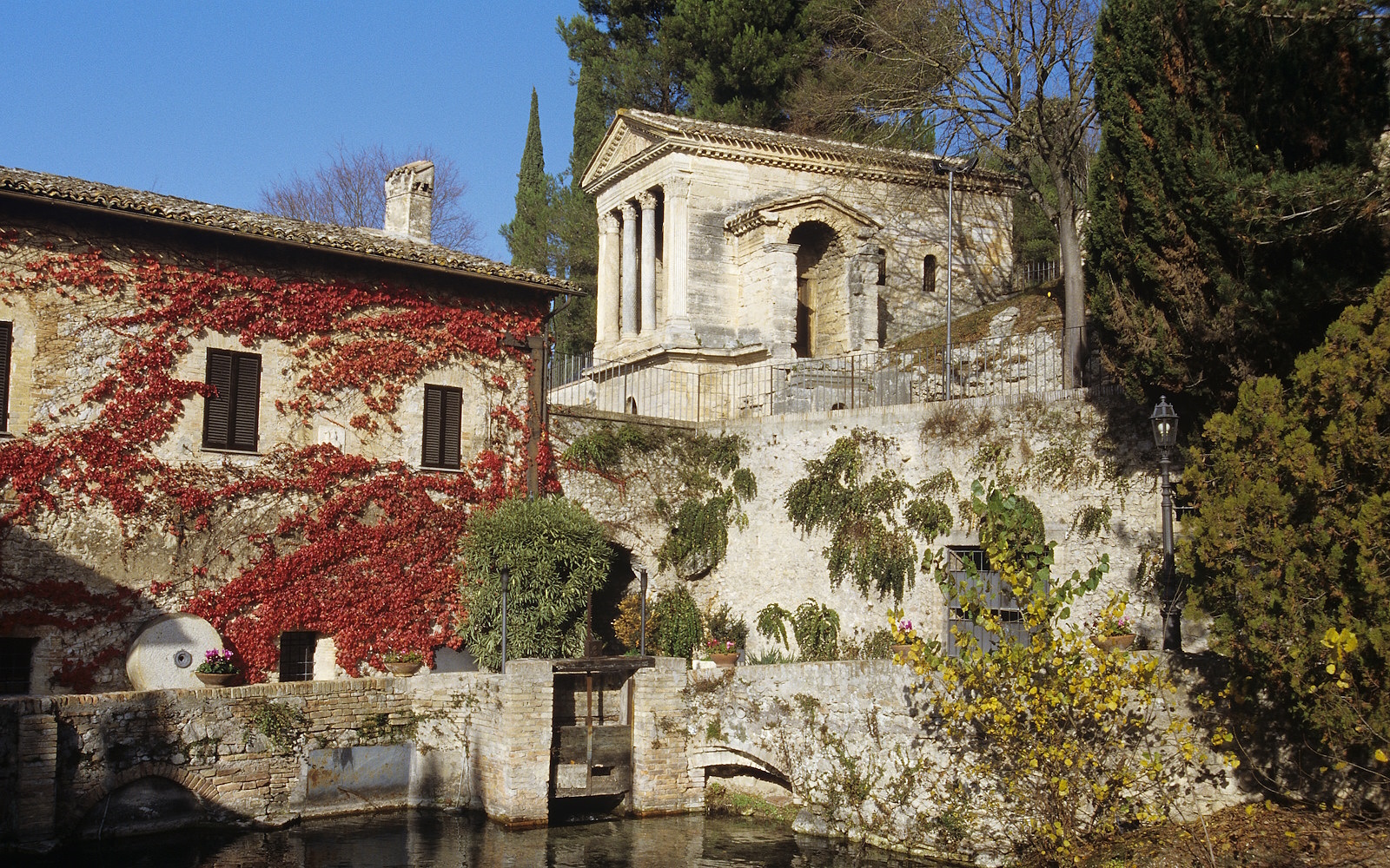 The Temple of Clitunno standing above and behind an ivy-covered building next to a canal
