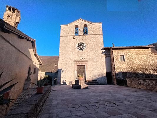 Vista frontale della Chiesa di San Giovanni a Vallo di Nera affacciata sulla piazza del borgo, con pozzo al centro ed edifici storici ai lati