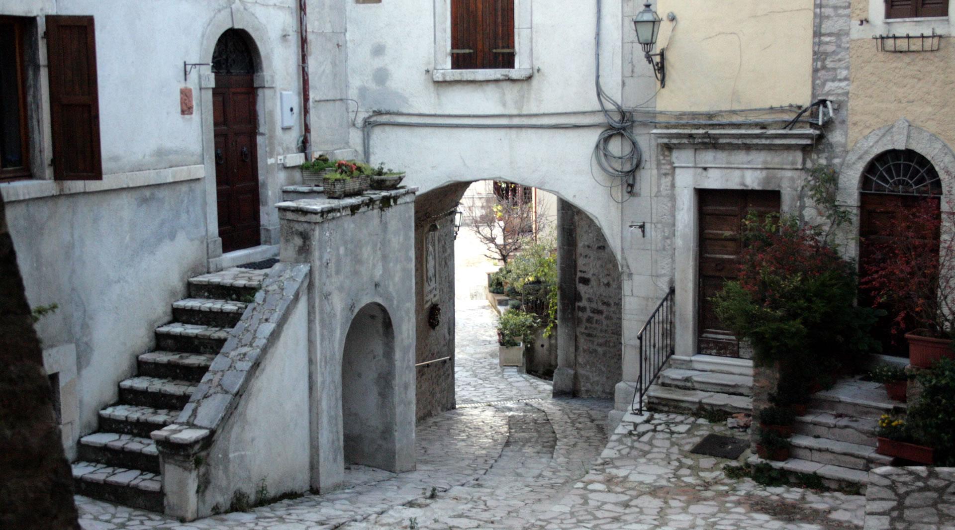 Une ruelle en pierre avec des arcs, des escaliers et des maisons historiques donnant sur une place pavée en pierre.