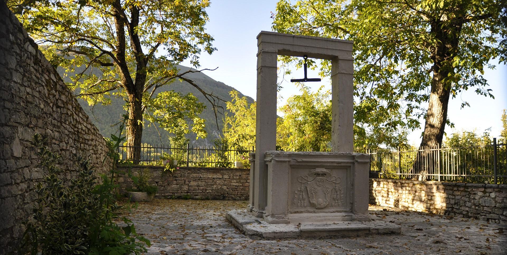 Puits historique en pierre avec architrave et armoiries de la famille Poli, entouré d’arbres et de murs à Usigni, Poggiodomo.