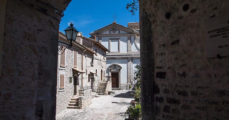 Vue de l’église San Salvatore à Usigni à travers une arche en pierre, avec des maisons historiques et une rue pavée.