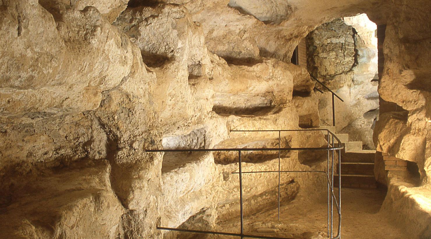 Ingresso in un corridoio sotterraneo della catacomba di Villa San Faustino. In primo piano, una scala stretta con un corrimano in ferro scende verso un lungo corridoio, dove si possono osservare loculi e nicchie scavati nelle pareti di mattone e pietra.