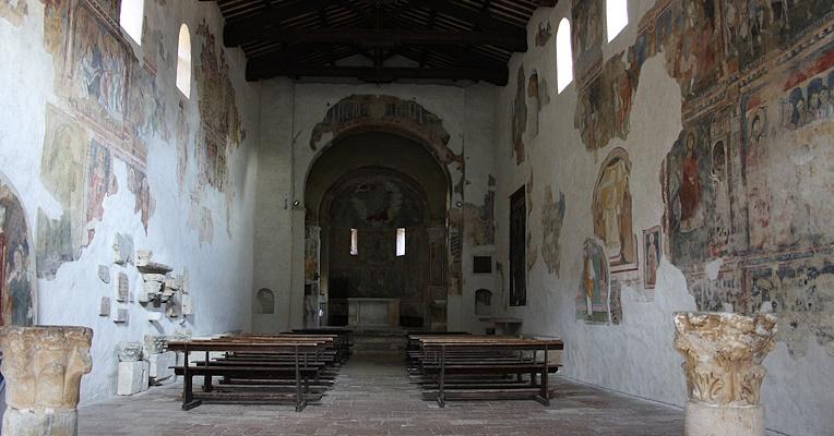 Interior of the Abbey of San Pietro in Valle in Ferentillo, with a view of the central nave and the apse. The walls feature medieval frescoes and arched windows, while the nave is flanked by wooden benches and the ceiling is made of exposed wooden beams.