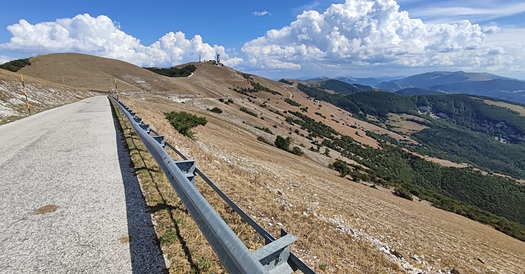 Mountain scenic road among barren hills and green valleys, with a blue sky and towering clouds on the horizon.