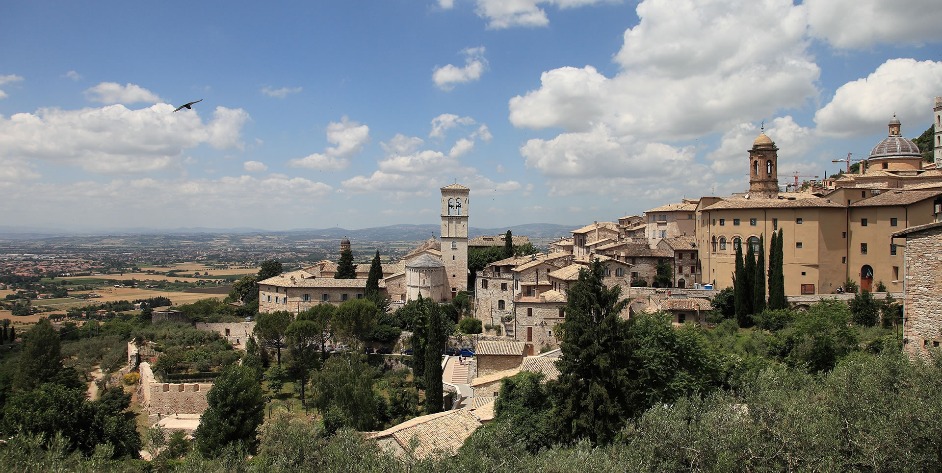 Panorama of Assisi with historic stone buildings, bell towers and towers under a clear sky but with some clouds