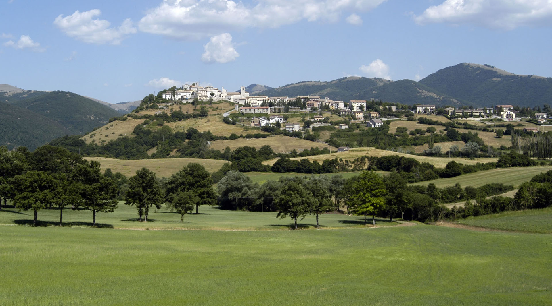 Panorama of Monteleone di Spoleto: the village sits on a green hill, framed by woods and Apennine mountains.