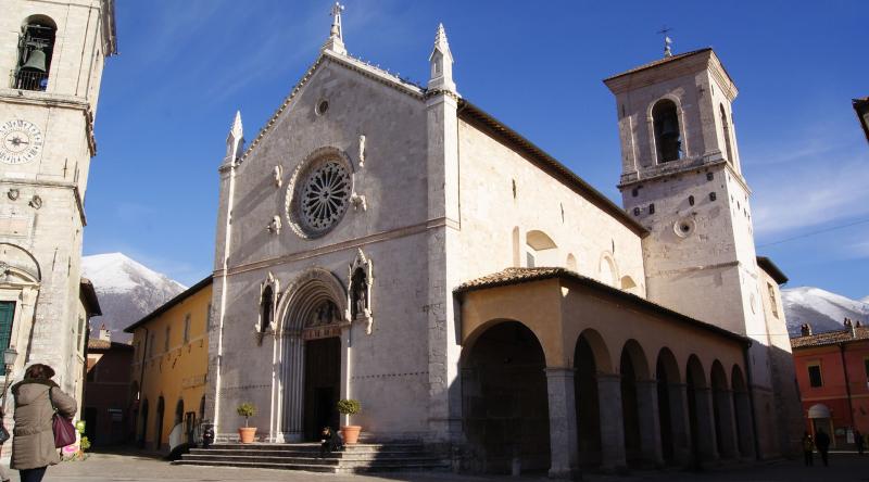 Basilika des Heiligen Benedikt in Norcia mit ihrer gotischen Fassade und Rosette, umrahmt von schneebedeckten Bergen unter klarem Himmel.