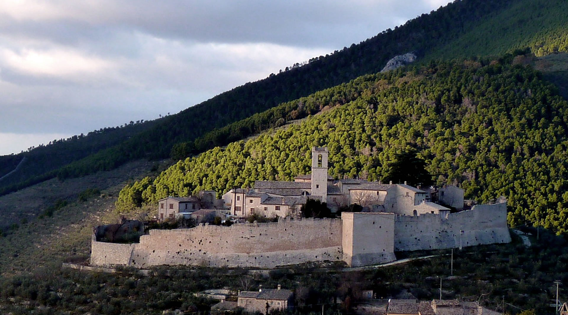 Medieval castle of Campello Alto, with surrounding walls and a bell tower lit by the sun, surrounded by mountains