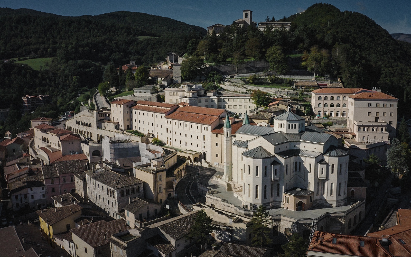 Aerial view of the Basilica of Saint Rita of Cascia complex, with red roofs and domes, nestled among Umbrian hills.