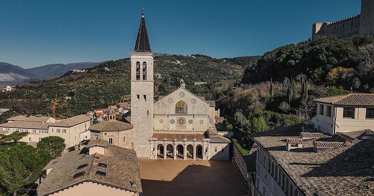 Aerial view of Spoleto Cathedral with Romanesque façade, bell tower and square, surrounded by hills and historic rooftops.