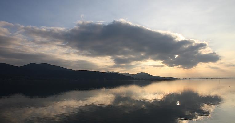 Une vue pittoresque du lac Trasimène au coucher du soleil, avec des nuages se reflétant sur l’eau calme et des montagnes en arrière-plan.