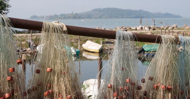 Reti da pesca con galleggianti appese a un palo, con vista sul lago Trasimeno, barche ormeggiate e un'isola sullo sfondo.