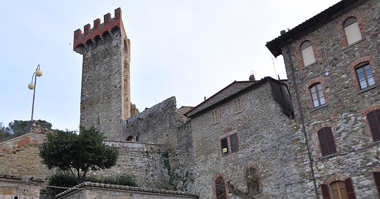 Medieval stone tower with red battlements, surrounded by stone buildings with arched windows and brick details.