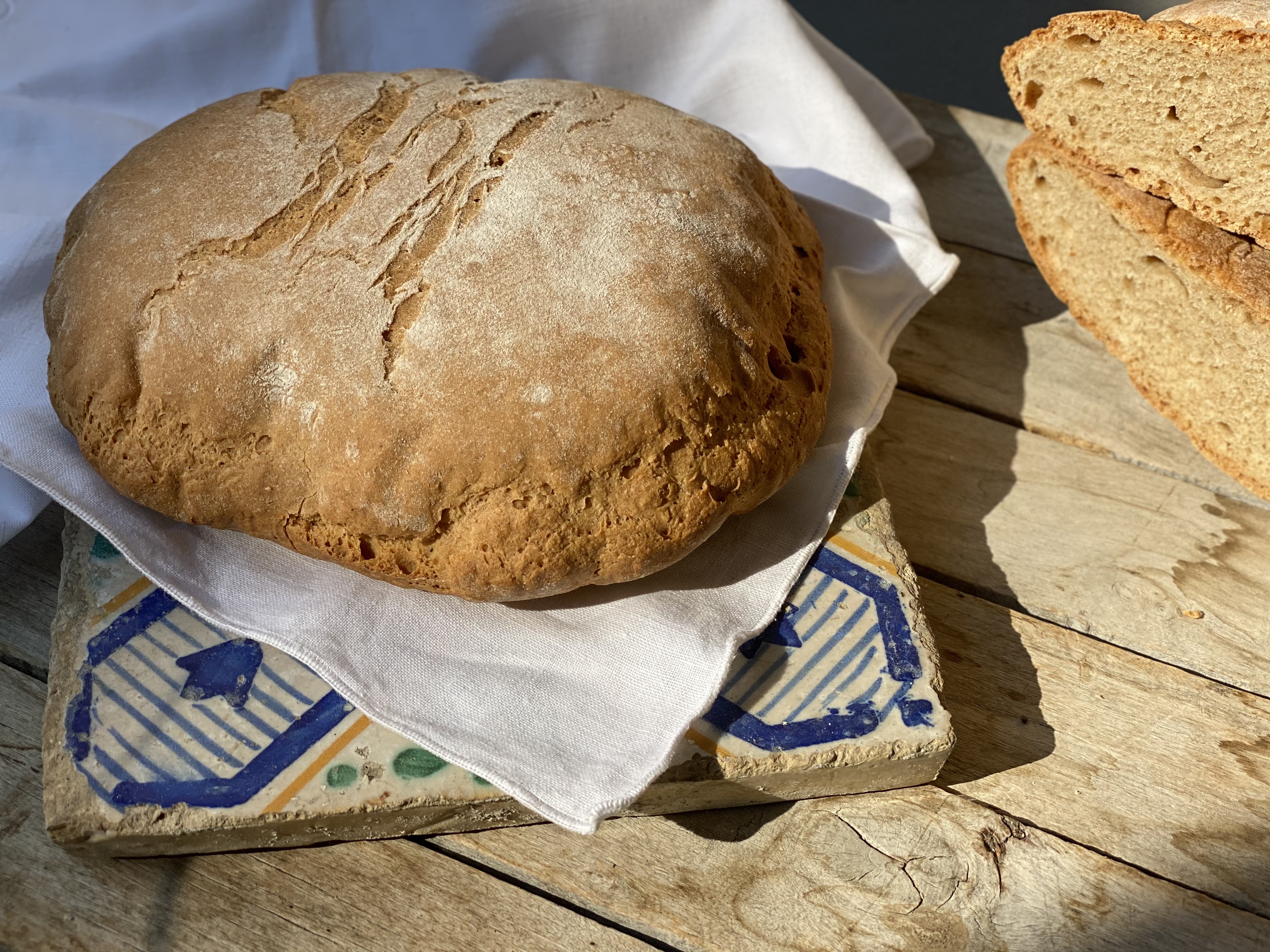 Rustic spelt bread roll on a decorated tile, with slices of bread beside it, lit by natural light.
