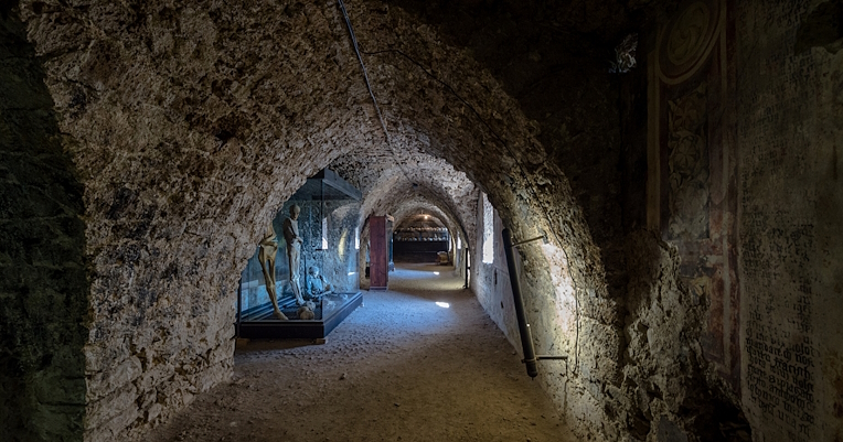 An underground corridor with stone walls, display cases containing mummies, and faded ancient frescoes.