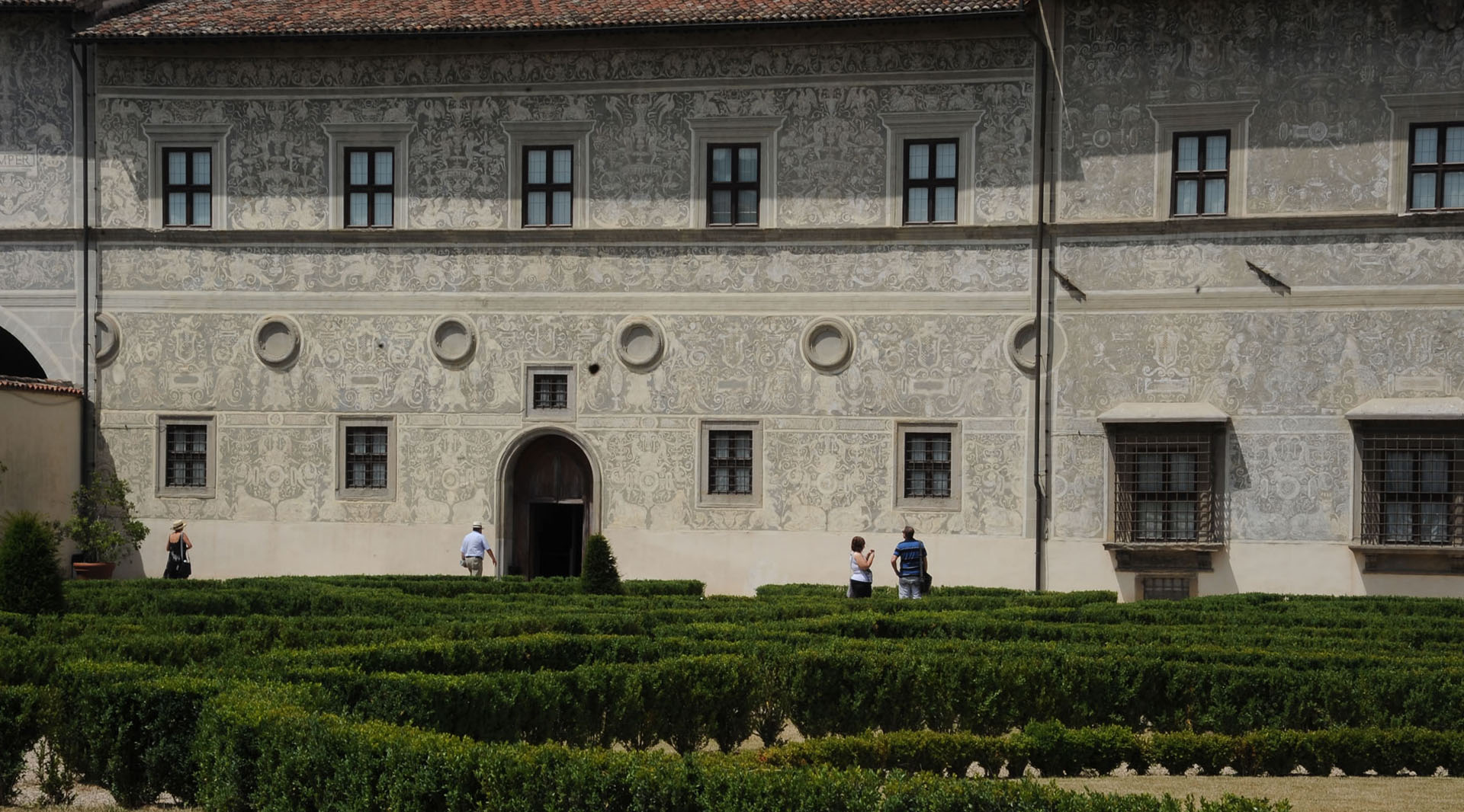 Palastfassade mit Sgraffitodekoren, rechteckigen Fenstern und einem Garten mit niedrigen Hecken.