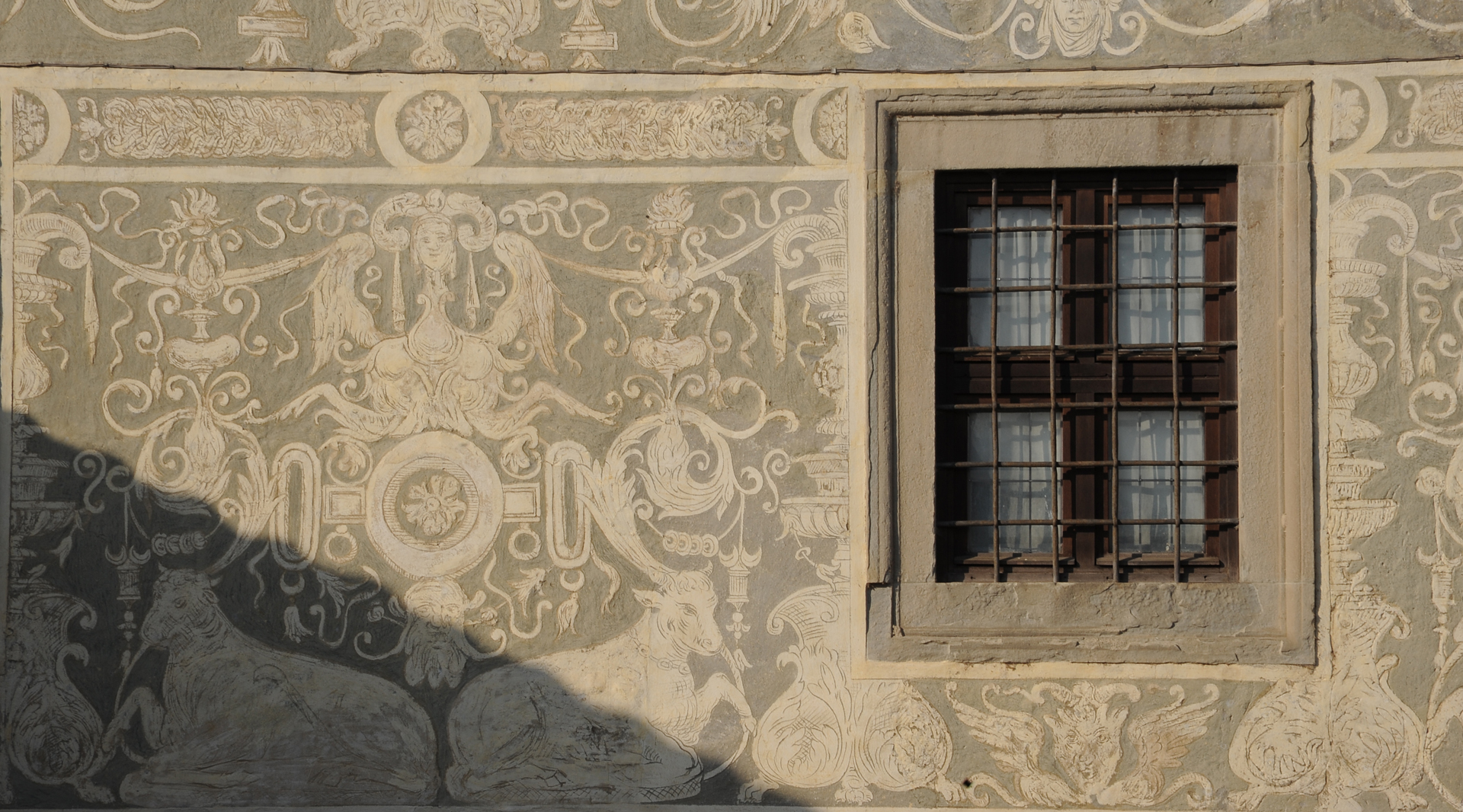 Fassadendetail mit Sgraffitodekoren und einem Fenster mit Eisengitter und Steinumrahmung.