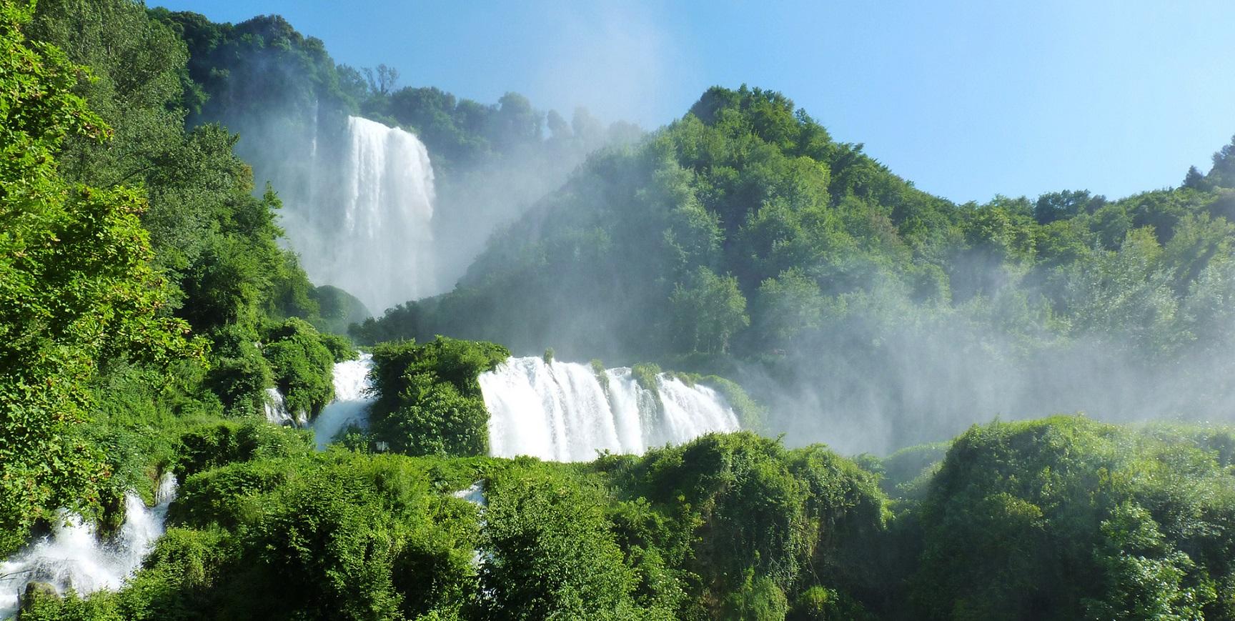 The Marmore Waterfall seen from below: cascades of water among dense vegetation, with spray and mist rising towards the clear sky.