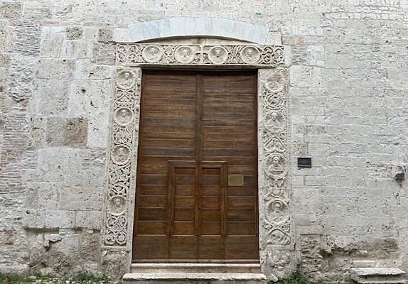 Porta in legno dell'ex chiesa di San Domenico a Narni, ora Auditorium Bortolotti, ornata con una cornice marmorea che presenta una serie di medaglioni scolpiti con ritratti di figure religiose. Sopra la porta, un arco in pietra bianca completa l'entrata monumentale.