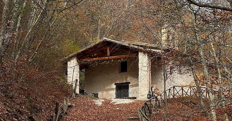 Small stone hermitage with a bell tower and wooden porch, nestled among trees on a mountain slope.