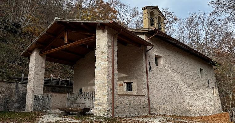 Small stone hermitage with a bell tower and wooden porch, nestled among trees on a mountain slope.