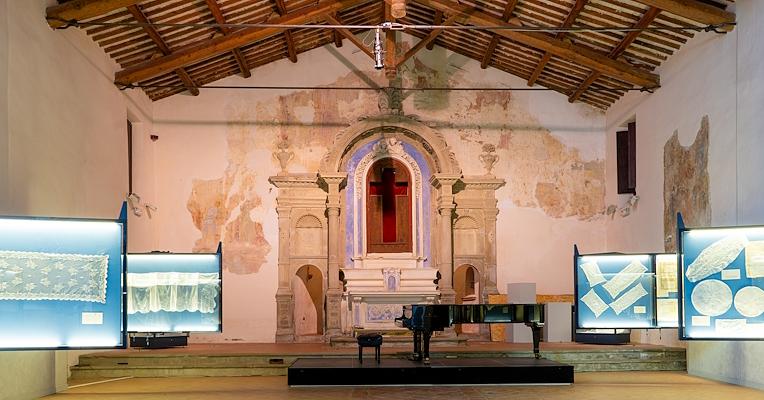 Interno della chiesa ora sede del museo, con vetrine espositive di merletti e un altare sullo sfondo. Al centro un pianoforte.