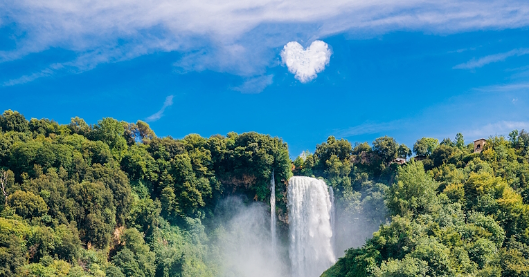 Cascata delle Marmore con una leggera nebbiolina e circondata dalla vegetazione. Nel cielo, una nuvola a forma di cuore