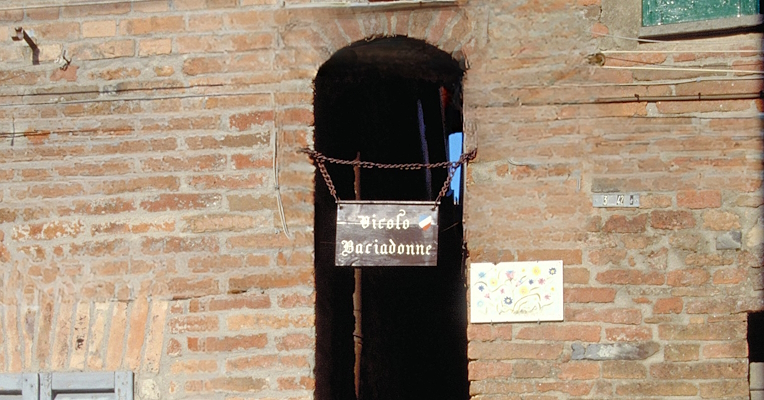 Stretto vicolo tra mura di mattoni, con un cartello marrone appeso a catene con la scritta bianca: “Vicolo Baciadonne”