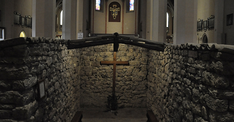 Interior of the Sanctuary of the Sacred Tugurio of Rivotorto, with stone walls, a wooden crucifix and a church in the background