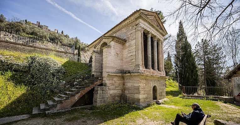 Exterior view of the Temple of Clitumnus, with four columns and a side staircase. In the foreground, a man sitting on a bench