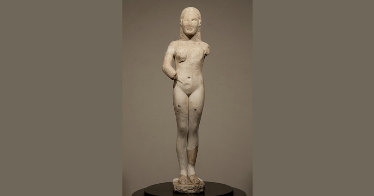 Statua arcaica femminile in marmo bianco, con parti mancanti e restaurate, in posa frontale.