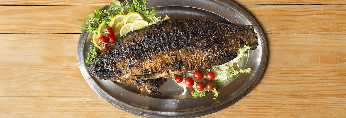 Carp in porchetta on a steel tray, garnished with lemon, salad, and cherry tomatoes on a wooden table.