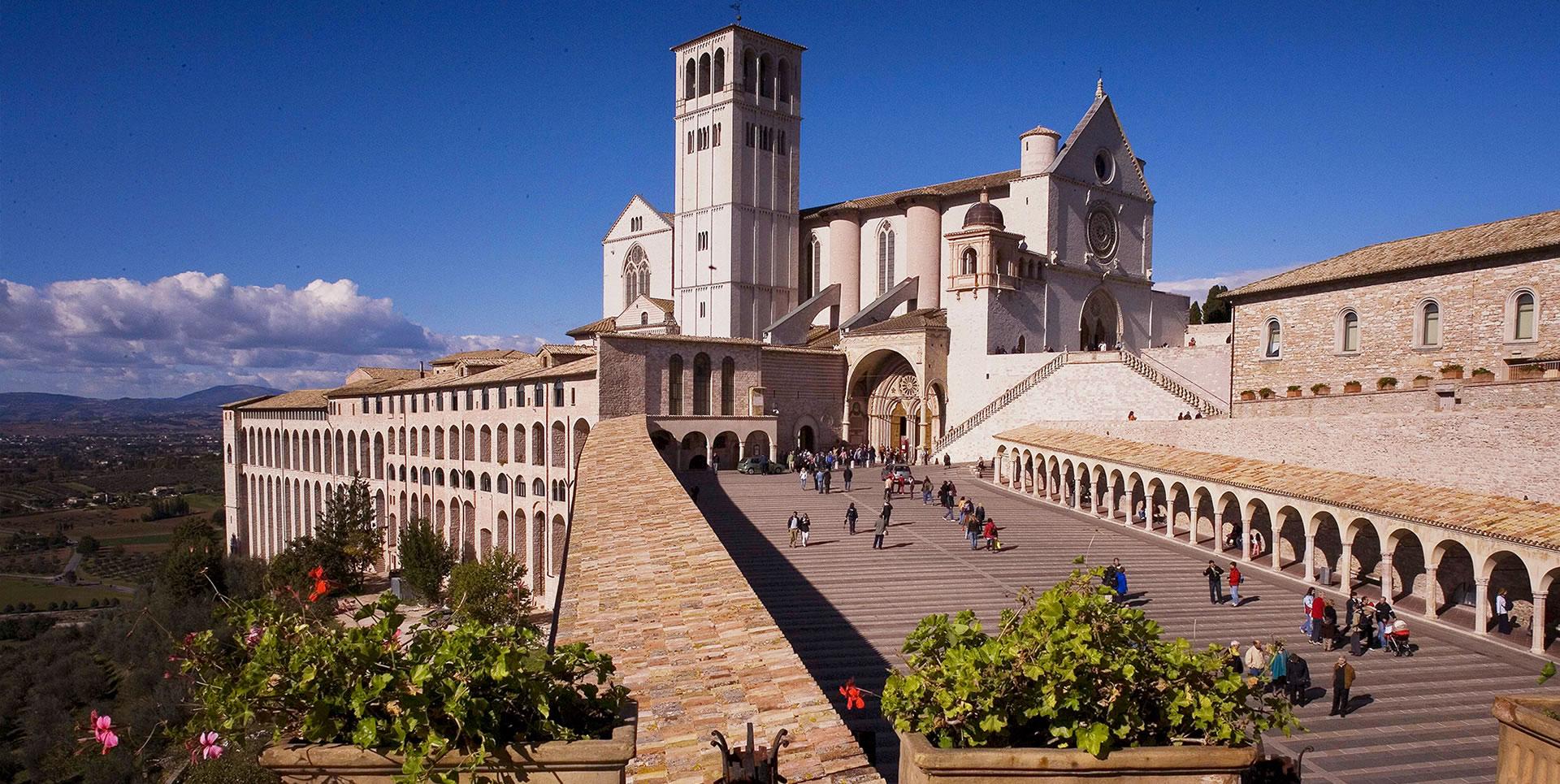 Panoramic view of the Basilica of St Francis in Assisi, showing the Upper Basilica in Gothic style, the large Lower Square with its side porticoes, and the central staircase leading to the entrance.