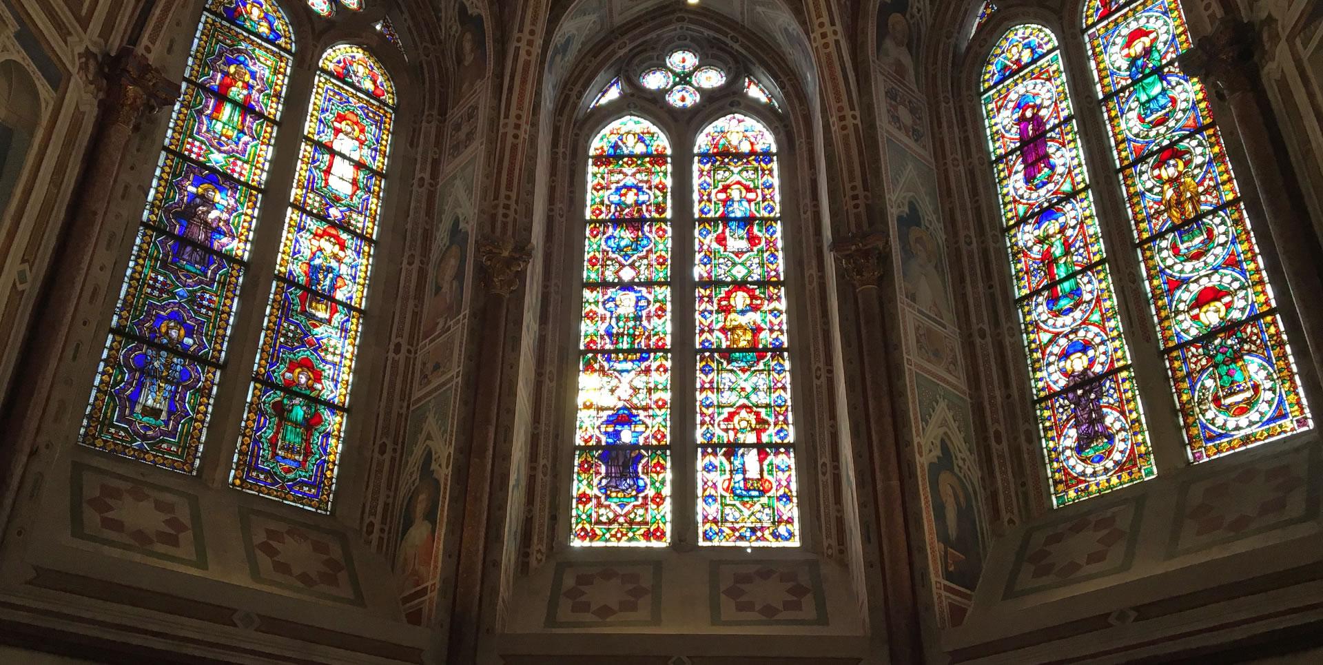 Stained glass windows inside the Basilica of St Francis in Assisi, with coloured details illustrating biblical scenes and figures of saints. The stained glass windows are positioned in the apse of the basilica and illuminate the interior.