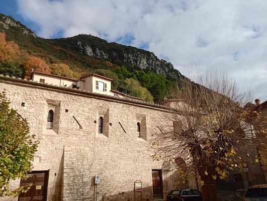 Stone building with arched windows in a village backed by tall mountains, with autumn trees and a partly cloudy sky.
