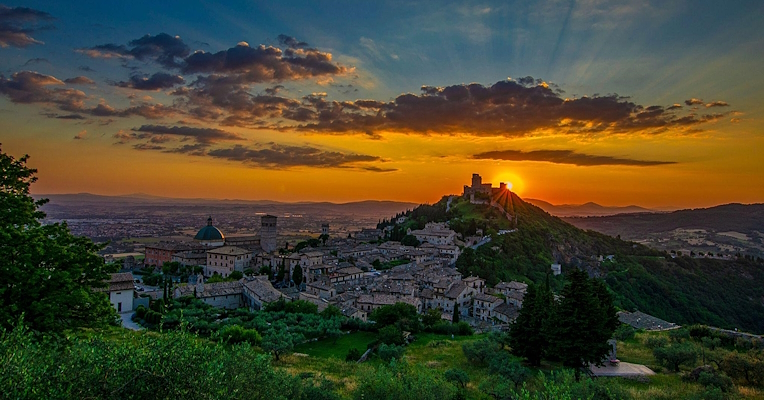 Vue panoramique d'Assise au coucher du soleil, avec le soleil se cachant derrière une colline et un ciel teinté d'orange et de bleu.