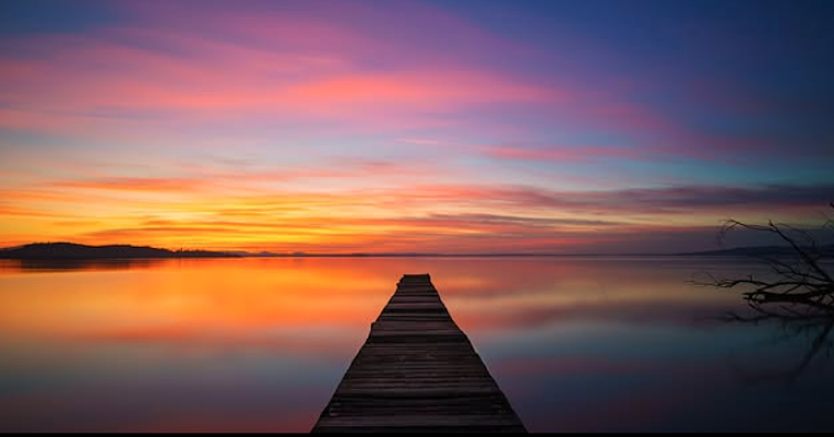 Jetée en bois sur les eaux du lac Trasimène, avec un ciel au coucher du soleil aux nuances de bleu, violet et orange reflété sur l’eau.