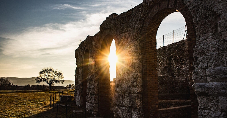 Soleil couchant derrière une arche du théâtre romain de Gubbio, créant un jeu de lumière chaude et d’ombres sur le paysage environnant.