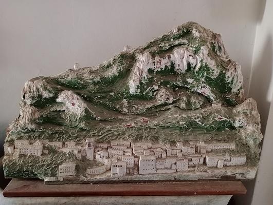 Plastico tridimensionale di un borgo ai piedi di una montagna, realizzato in gesso dipinto con edifici bianchi e rilievi verdi.