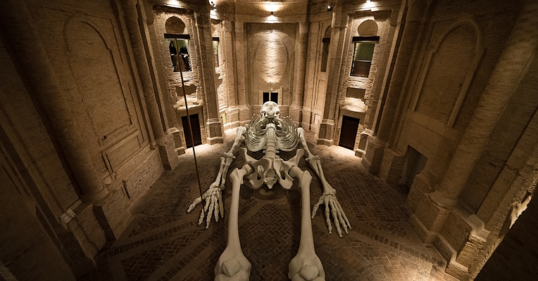 Installazione artistica di uno scheletro gigante disteso all'interno di una cappella con architettura in mattoni.