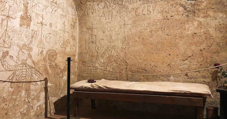 Walls engraved with symbols, crosses, and centuries-old inscriptions in a prison cell lit by a small barred window.