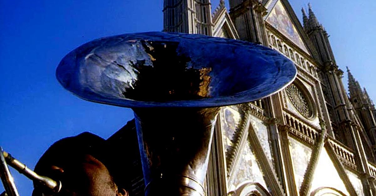 Man playing a tuba, seen from below, with the façade of Orvieto Cathedral and a clear blue sky in the background.