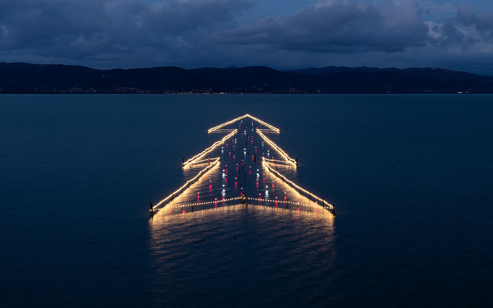 Illuminated floating Christmas Tree on Lake Trasimeno at sunset, reflected in the calm waters.