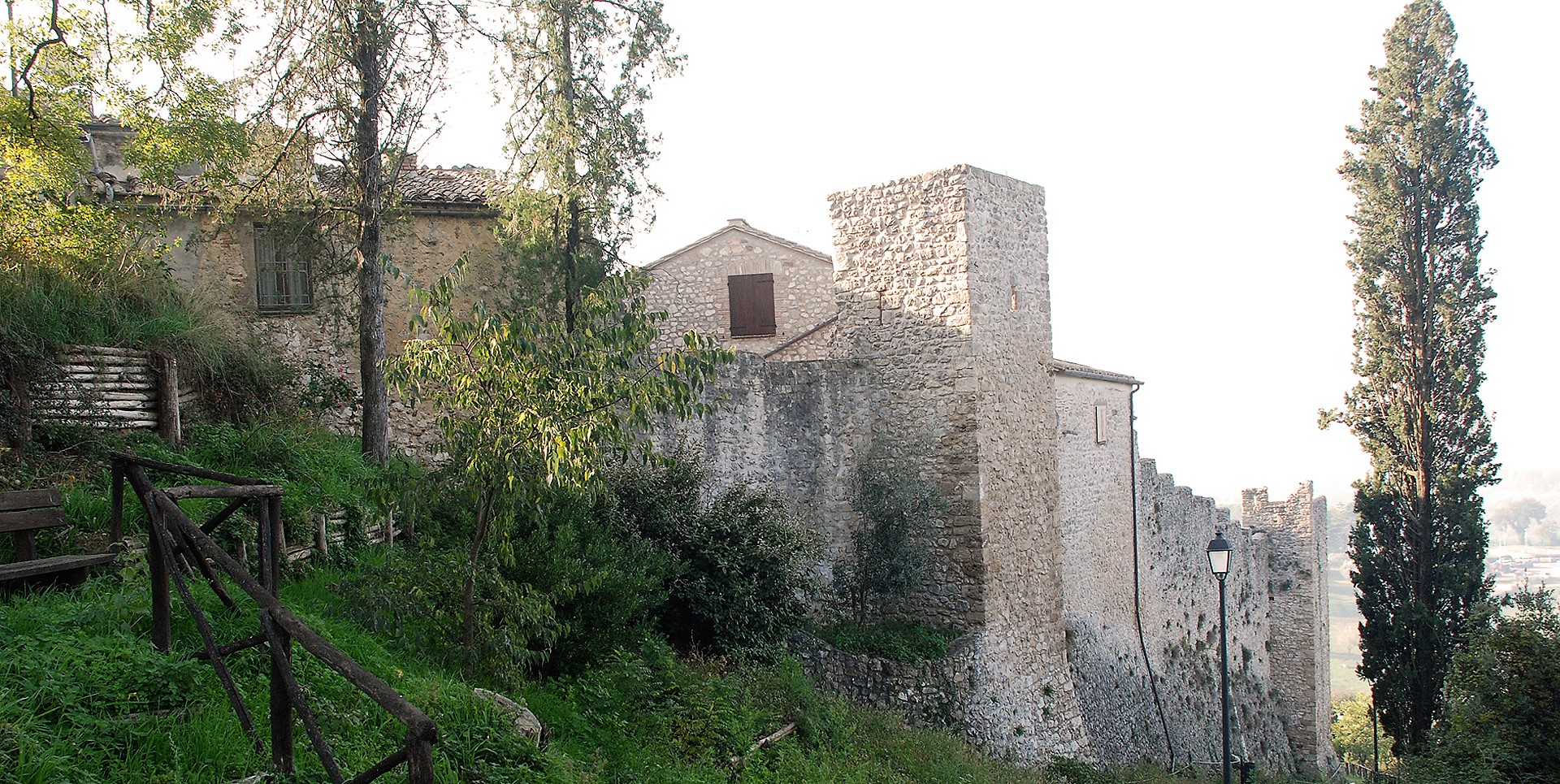 Vue latérale du château de Poggio di Otricoli avec ses murs en pierre, sa tour carrée et la végétation le long de la pente.